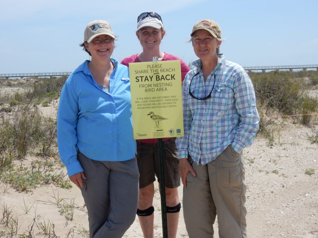 three smiling volunteers stand with a beach nesting birds sign